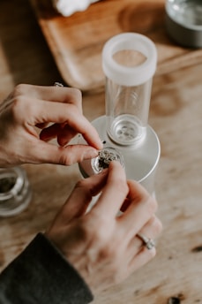 A person's hands are placing a small amount of dried herbal material into a glass chamber on a metallic device. In the background, there is a wooden tray and some other objects slightly out of focus.