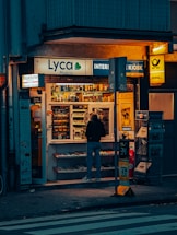 A small kiosk is brightly illuminated against the dim evening surroundings. The kiosk's shelves are filled with a variety of goods like snacks, magazines, and lottery tickets. A person stands in front, browsing the items inside. Advertisements and signage from brands like Lyca Mobile and DHL are visible, contributing to the urban character of the scene.