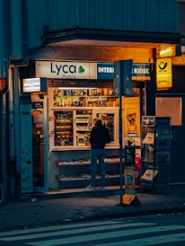 A small kiosk is brightly illuminated against the dim evening surroundings. The kiosk's shelves are filled with a variety of goods like snacks, magazines, and lottery tickets. A person stands in front, browsing the items inside. Advertisements and signage from brands like Lyca Mobile and DHL are visible, contributing to the urban character of the scene.
