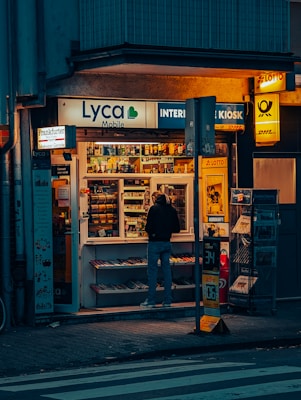 A small kiosk is brightly illuminated against the dim evening surroundings. The kiosk's shelves are filled with a variety of goods like snacks, magazines, and lottery tickets. A person stands in front, browsing the items inside. Advertisements and signage from brands like Lyca Mobile and DHL are visible, contributing to the urban character of the scene.