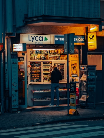 A small kiosk is brightly illuminated against the dim evening surroundings. The kiosk's shelves are filled with a variety of goods like snacks, magazines, and lottery tickets. A person stands in front, browsing the items inside. Advertisements and signage from brands like Lyca Mobile and DHL are visible, contributing to the urban character of the scene.