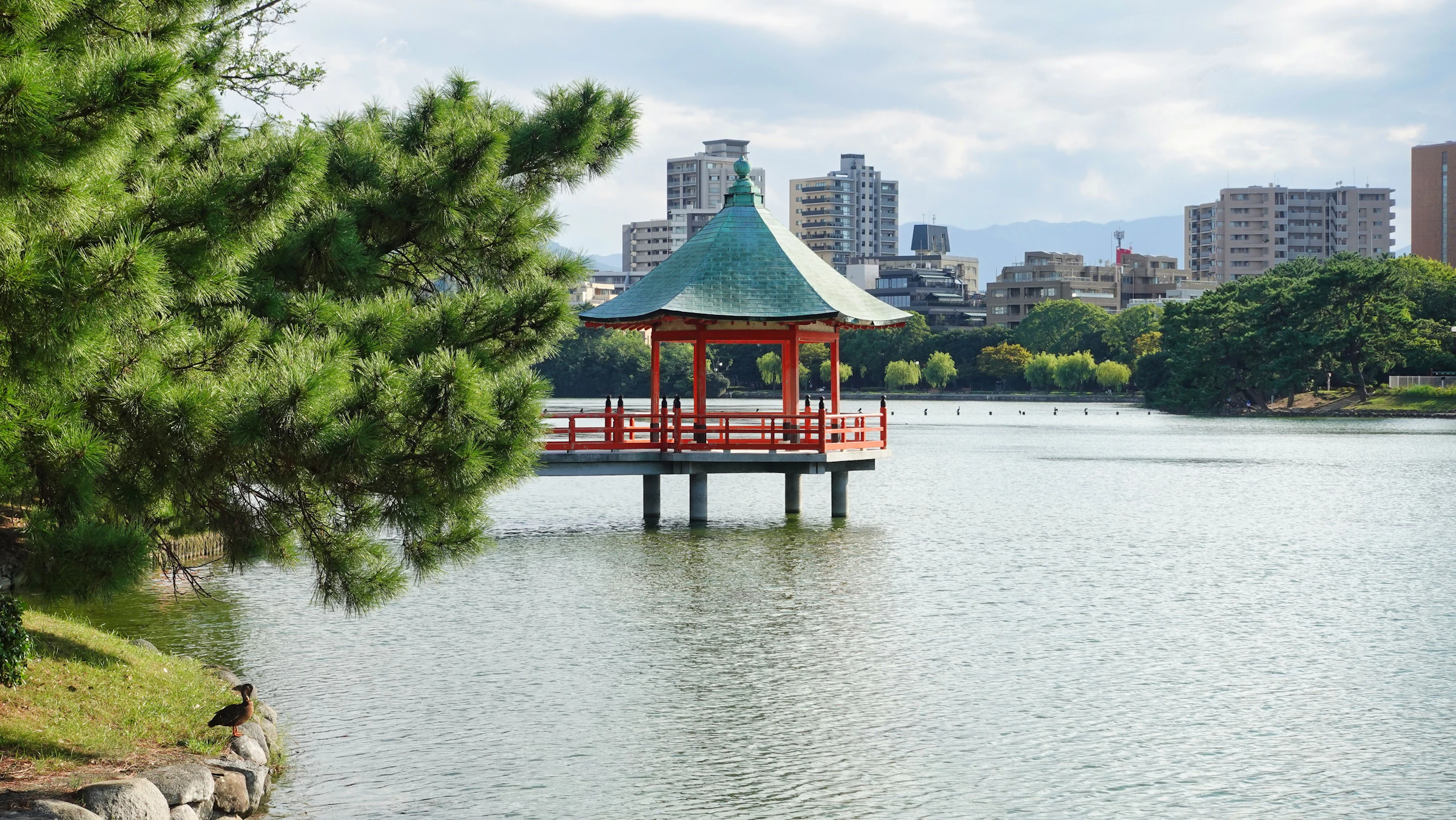 A red pavilion on the lake at Ohori Park with the Fukuoka skyline behind it
