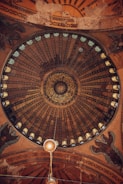An intricately designed dome featuring concentric circles with detailed geometric and calligraphic patterns. The dome includes motifs of eagles situated symmetrically with a rich palette of earthy colors. A central light fixture hangs from the middle of the dome.