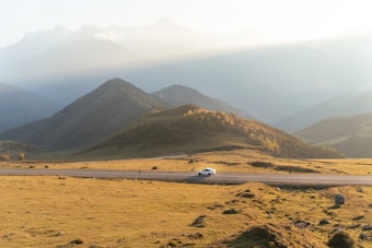 A white car travels on a road set against a backdrop of rolling hills and mountains. The landscape is illuminated by soft, golden sunlight that casts gentle shadows over the grassy terrain. The mountains in the distance are shrouded in mist, adding depth and a serene atmosphere to the scene.