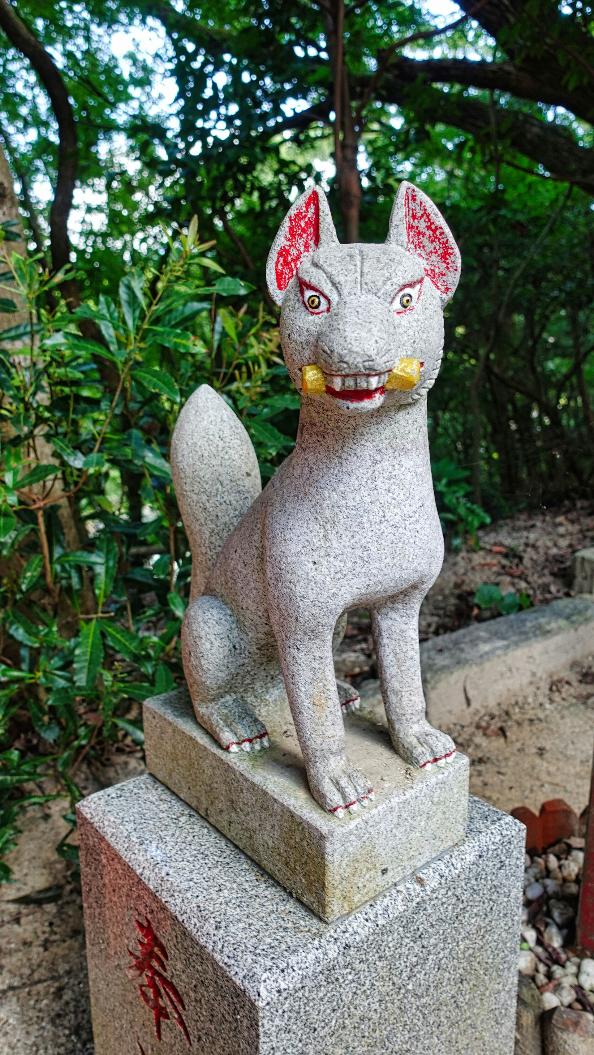 A stone statue of a fox with detailed features stands on a pedestal. The fox has a striking expression with gold elements in its mouth and vibrant red inside its ears. It is surrounded by lush green foliage, suggesting a natural or garden setting.