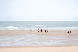 A group of people in swimwear walks along a wide, flat beach with gentle waves from the sea. The sky is clear, and the sand is smooth with patches of wet areas, reflecting a soft, natural ambiance.