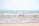 A group of people in swimwear walks along a wide, flat beach with gentle waves from the sea. The sky is clear, and the sand is smooth with patches of wet areas, reflecting a soft, natural ambiance.