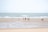 A group of people in swimwear walks along a wide, flat beach with gentle waves from the sea. The sky is clear, and the sand is smooth with patches of wet areas, reflecting a soft, natural ambiance.