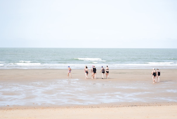 A group of people in swimwear walks along a wide, flat beach with gentle waves from the sea. The sky is clear, and the sand is smooth with patches of wet areas, reflecting a soft, natural ambiance.