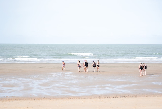 A group of people in swimwear walks along a wide, flat beach with gentle waves from the sea. The sky is clear, and the sand is smooth with patches of wet areas, reflecting a soft, natural ambiance.