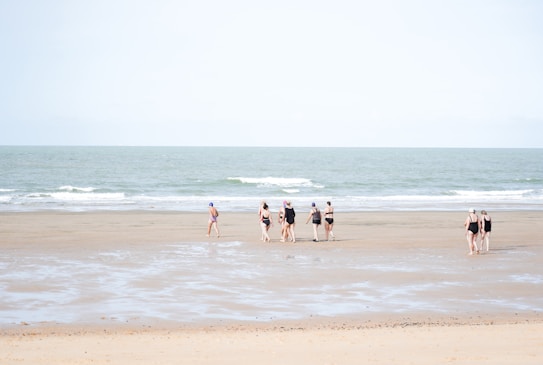 A group of people in swimwear walks along a wide, flat beach with gentle waves from the sea. The sky is clear, and the sand is smooth with patches of wet areas, reflecting a soft, natural ambiance.