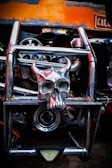 A metallic skull with horns is mounted on a chrome vehicle grill, surrounded by gears and mechanical parts. The background includes orange and black colors, with a visible plate on the top right corner.