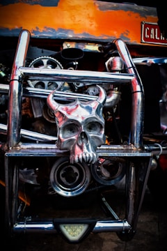 A metallic skull with horns is mounted on a chrome vehicle grill, surrounded by gears and mechanical parts. The background includes orange and black colors, with a visible plate on the top right corner.