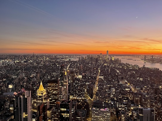 A stunning cityscape at sunset, with a sprawling view of skyscrapers and a beautifully colored sky transitioning from blue to warm orange hues. Streets illuminated with lights can be seen below, and a river is visible in the background.