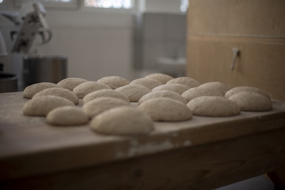 Several pieces of dough are neatly arranged on a wooden table in a kitchen setting, preparing them for baking. In the background, blurred elements of a mixing appliance can be seen, suggesting a professional or home-like baking environment.