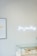 A minimalistic jewelry store display featuring a glass case filled with various items like rings and necklaces. Above the case, a bright neon sign spells 'fine jewellery'. The wall is lined with vertical white wooden panels, and a framed picture is partially visible, showing stacked jewelry.