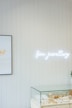 A minimalistic jewelry store display featuring a glass case filled with various items like rings and necklaces. Above the case, a bright neon sign spells 'fine jewellery'. The wall is lined with vertical white wooden panels, and a framed picture is partially visible, showing stacked jewelry.