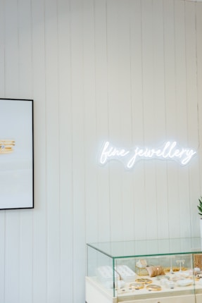 A minimalistic jewelry store display featuring a glass case filled with various items like rings and necklaces. Above the case, a bright neon sign spells 'fine jewellery'. The wall is lined with vertical white wooden panels, and a framed picture is partially visible, showing stacked jewelry.