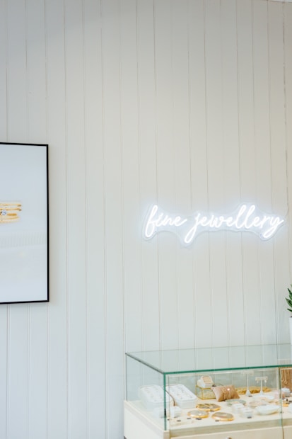 A minimalistic jewelry store display featuring a glass case filled with various items like rings and necklaces. Above the case, a bright neon sign spells 'fine jewellery'. The wall is lined with vertical white wooden panels, and a framed picture is partially visible, showing stacked jewelry.