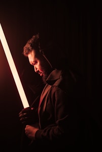 A person in a dark environment holding a glowing orange lightsaber-like object. The individual is wearing a dark hooded garment, and their face is illuminated by the warm glow. The overall ambiance is mysterious and dramatic, with strong contrasts between the light and shadows.