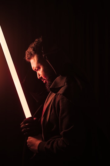 A person in a dark environment holding a glowing orange lightsaber-like object. The individual is wearing a dark hooded garment, and their face is illuminated by the warm glow. The overall ambiance is mysterious and dramatic, with strong contrasts between the light and shadows.