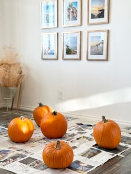 Five pumpkins are arranged on a wooden floor covered with newspaper. The pumpkins are bright orange and vary slightly in size. In the background, a white wall features six framed photographs or artworks, arranged in two rows. Near the corner, a bundle of dried plants complements the simple and cozy setting.
