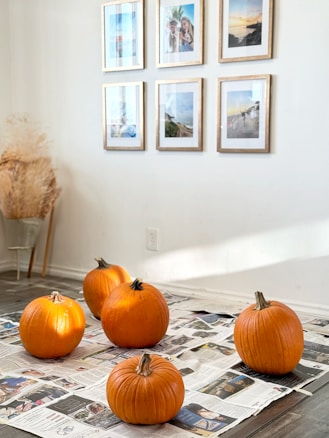 Five pumpkins are arranged on a wooden floor covered with newspaper. The pumpkins are bright orange and vary slightly in size. In the background, a white wall features six framed photographs or artworks, arranged in two rows. Near the corner, a bundle of dried plants complements the simple and cozy setting.