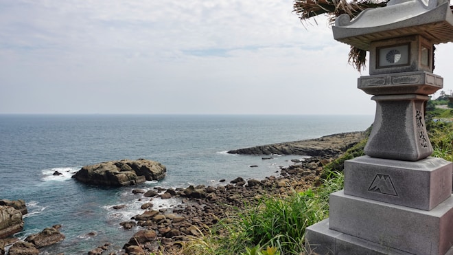 A serene coastal landscape with rocky formations extending into the calm sea under a slightly overcast sky. A traditional stone lantern is prominently positioned on the right side, partially surrounded by green foliage.