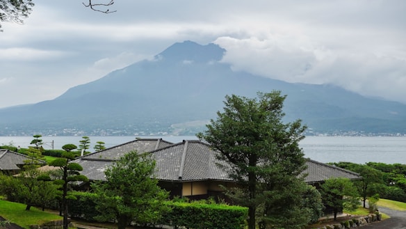 A tranquil landscape featuring a traditional Japanese house with a gray tiled roof surrounded by lush green trees. In the background, a large mountain is partially covered by clouds, with a calm body of water separating the house from the mountain. The scene conveys a peaceful and harmonious connection with nature.