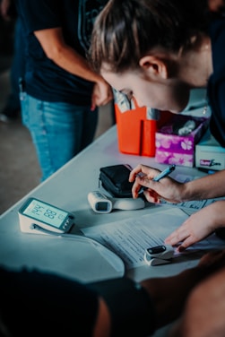 A person is leaning over a table, writing on papers with a pen. There are medical devices, including a blood pressure monitor and a pulse oximeter, on the table. The setting appears to be an informal medical or health-related event, with another person partially visible in the background.