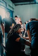 A group of people are gathered indoors, with one individual sitting and having their blood pressure checked. Others stand around engaging in various activities, suggesting a community or clinic setting. The lighting is calm and natural, with a mix of shadows and highlights.