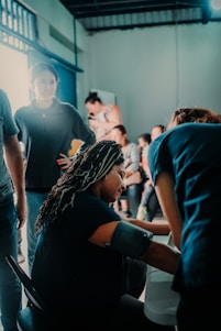 A group of people are gathered indoors, with one individual sitting and having their blood pressure checked. Others stand around engaging in various activities, suggesting a community or clinic setting. The lighting is calm and natural, with a mix of shadows and highlights.