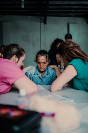A group of people gathered around a table, where a person in the center seems to be receiving comfort or support. The atmosphere appears intimate and focused, with individuals displaying attentive body language.