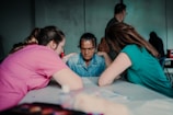 Three individuals are sitting around a table, deeply engaged in conversation. The central figure has a contemplative expression, while the other two, positioned on either side, appear supportive and attentive.