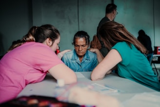 Three individuals are sitting around a table, deeply engaged in conversation. The central figure has a contemplative expression, while the other two, positioned on either side, appear supportive and attentive.