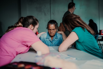 Three individuals are sitting around a table, deeply engaged in conversation. The central figure has a contemplative expression, while the other two, positioned on either side, appear supportive and attentive.