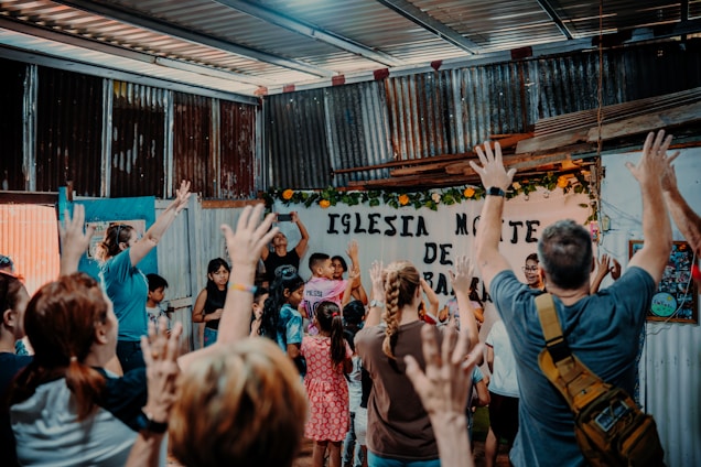 A group of people, including children and adults, gather inside a building with corrugated metal walls, raising their hands in unison. The atmosphere appears lively and engaged, with some participants standing close together. The background features a sign with Spanish text, partially obscured by flowers and decorations.
