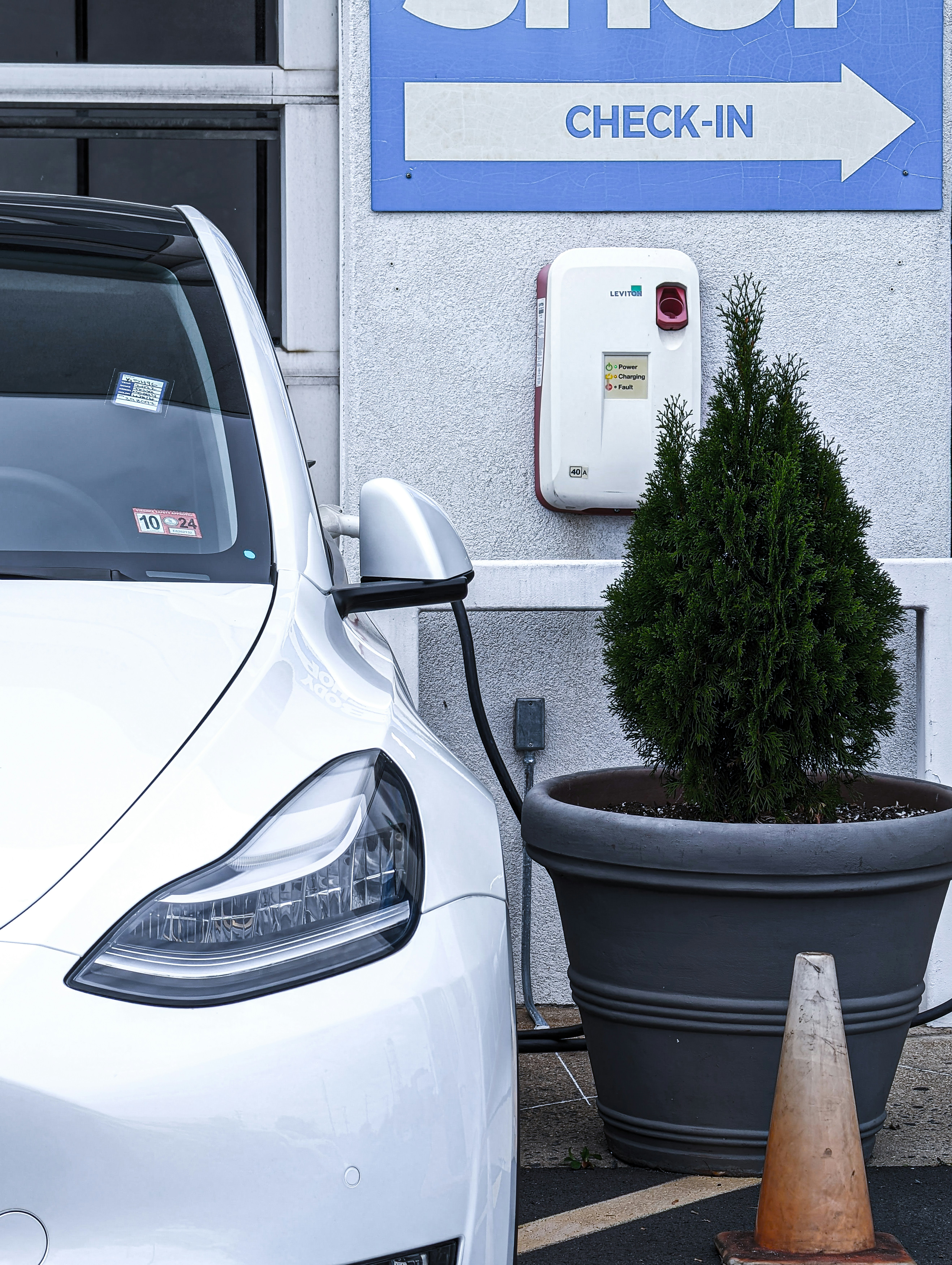 A white electric vehicle is plugged into a charging station, which is mounted on a light gray wall. To the right, a large planter containing a small coniferous tree is visible, with an orange traffic cone positioned next to it. Above, a sign with the word 'CHECK-IN' and an arrow points to the right.