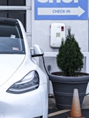 A white electric vehicle is plugged into a charging station, which is mounted on a light gray wall. To the right, a large planter containing a small coniferous tree is visible, with an orange traffic cone positioned next to it. Above, a sign with the word 'CHECK-IN' and an arrow points to the right.