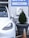 A white electric vehicle is plugged into a charging station, which is mounted on a light gray wall. To the right, a large planter containing a small coniferous tree is visible, with an orange traffic cone positioned next to it. Above, a sign with the word 'CHECK-IN' and an arrow points to the right.