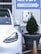 A white electric vehicle is plugged into a charging station, which is mounted on a light gray wall. To the right, a large planter containing a small coniferous tree is visible, with an orange traffic cone positioned next to it. Above, a sign with the word 'CHECK-IN' and an arrow points to the right.