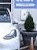 A white electric vehicle is plugged into a charging station, which is mounted on a light gray wall. To the right, a large planter containing a small coniferous tree is visible, with an orange traffic cone positioned next to it. Above, a sign with the word 'CHECK-IN' and an arrow points to the right.