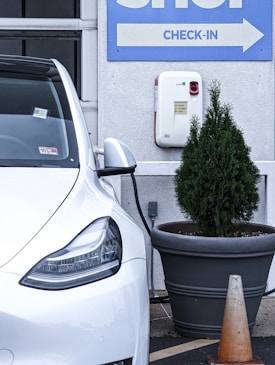 A white electric vehicle is plugged into a charging station, which is mounted on a light gray wall. To the right, a large planter containing a small coniferous tree is visible, with an orange traffic cone positioned next to it. Above, a sign with the word 'CHECK-IN' and an arrow points to the right.