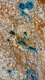 An aerial view showcasing a landscape with a mix of earthy brown tones and vibrant blue patches. The surface has a textured, rock-like appearance, with various shades and gradients blending together. The blue areas appear like water bodies or mineral deposits amidst the dry, rugged terrain.