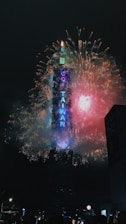 Bright and colorful fireworks surround a tall skyscraper, lighting up the night sky. The words on the building spell out 'GO TAIWAN,' illuminated in neon lighting.