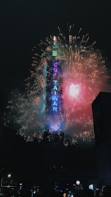 Bright and colorful fireworks surround a tall skyscraper, lighting up the night sky. The words on the building spell out 'GO TAIWAN,' illuminated in neon lighting.