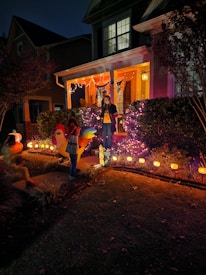 A house porch is decorated with Halloween-themed lights and ornaments. A person stands on the steps surrounded by purple and orange string lights, while two children in costumes are seen in the yard. Pumpkin decorations with glowing faces line the pathway.
