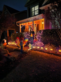 A house porch is decorated with Halloween-themed lights and ornaments. A person stands on the steps surrounded by purple and orange string lights, while two children in costumes are seen in the yard. Pumpkin decorations with glowing faces line the pathway.