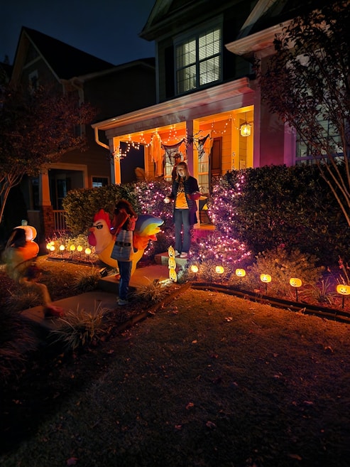 A house porch is decorated with Halloween-themed lights and ornaments. A person stands on the steps surrounded by purple and orange string lights, while two children in costumes are seen in the yard. Pumpkin decorations with glowing faces line the pathway.