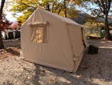 A large beige tent is set up in a leafy campsite surrounded by autumnal trees with colorful foliage. The ground is covered with small gravel stones and scattered fallen leaves. Two dark green canisters are placed near the entrance of the tent.